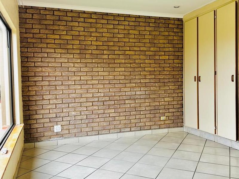 A bright, empty bedroom featuring a distinctive exposed brick feature wall, neutral tiled flooring, and ample built-in cupboard storage.