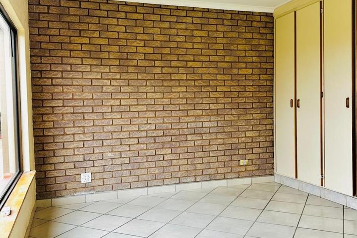 A bright, empty bedroom featuring a distinctive exposed brick feature wall, neutral tiled flooring, and ample built-in cupboard storage.