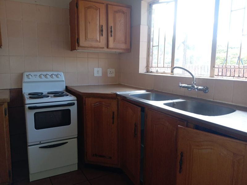 A dated kitchen featuring wooden cabinetry, a double basin sink, tiled backsplash, and a freestanding stove, with natural light from a window with security bars.