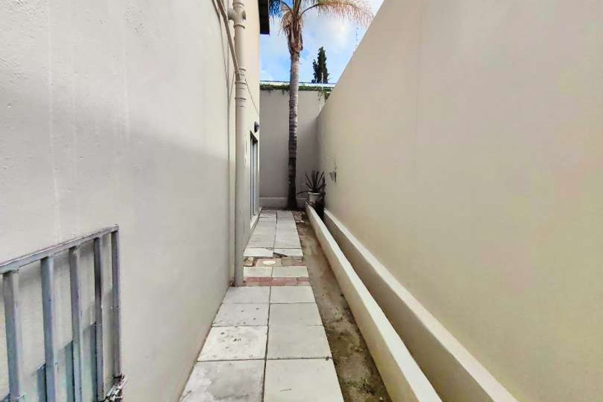 A narrow side passage featuring paved walkway, retaining wall, and exterior building access, requiring maintenance to the paving and ground surface.