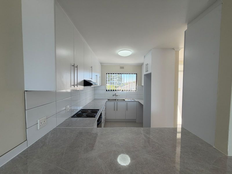 A clean, modern kitchen featuring white cabinetry, grey marble-effect countertops, an electric hob, and a double bowl sink with a window view.