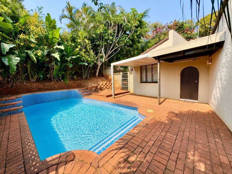 Outdoor pool area featuring a swimming pool, brick paved surround, and a covered patio space set against a lush garden backdrop.