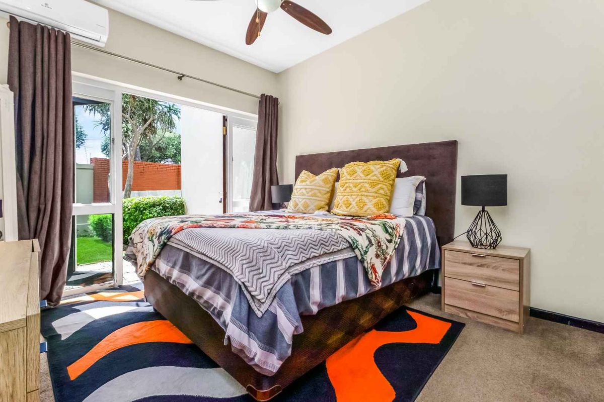 A bright bedroom featuring a ceiling fan, wall-mounted air conditioning unit, and large glass sliding doors providing direct access to the outdoor garden area.