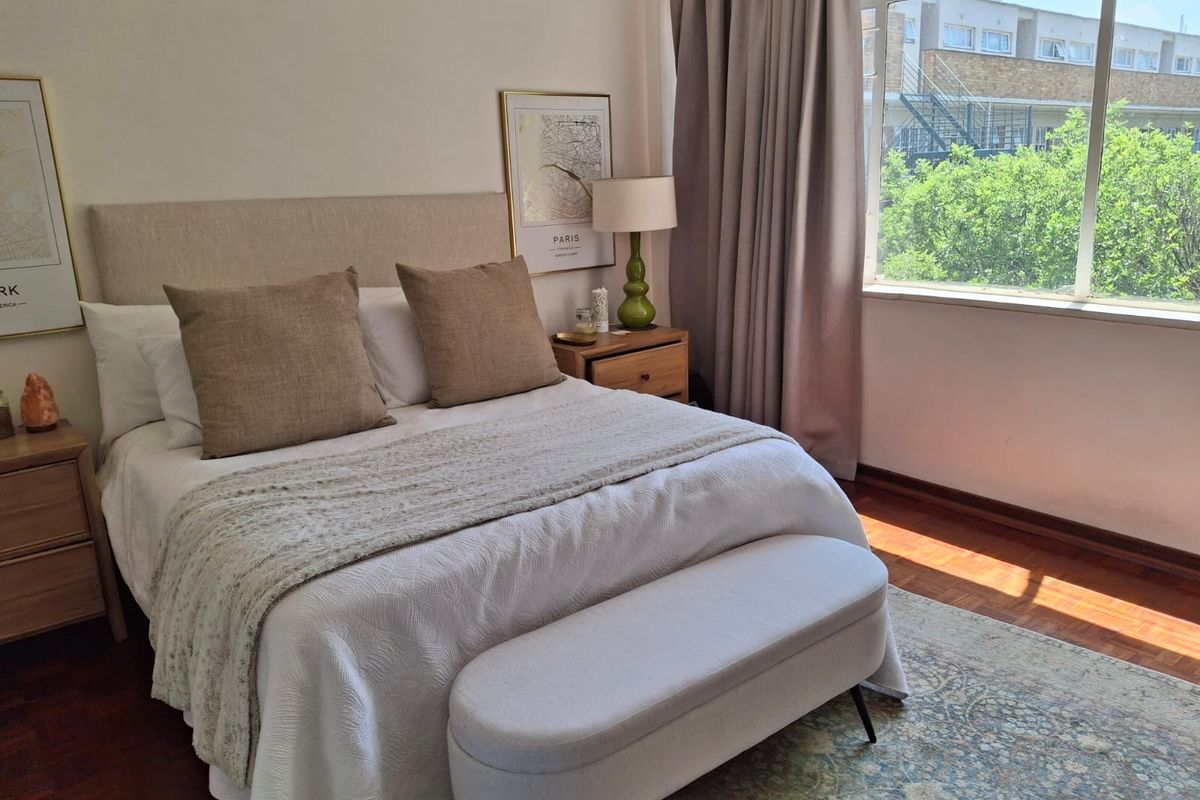 A well-lit bedroom featuring warm parquet flooring and a large window that provides natural light and a pleasant exterior view.