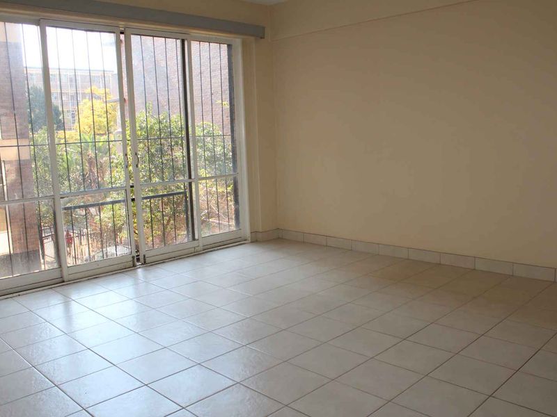 A bright, empty living area featuring neutral tiled flooring and a large sliding glass door with security bars, providing access to the exterior.
