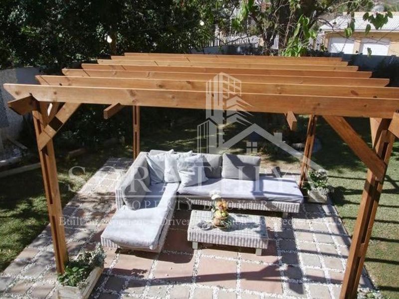 A well-maintained outdoor patio area featuring a sturdy wooden pergola structure set within a landscaped garden.