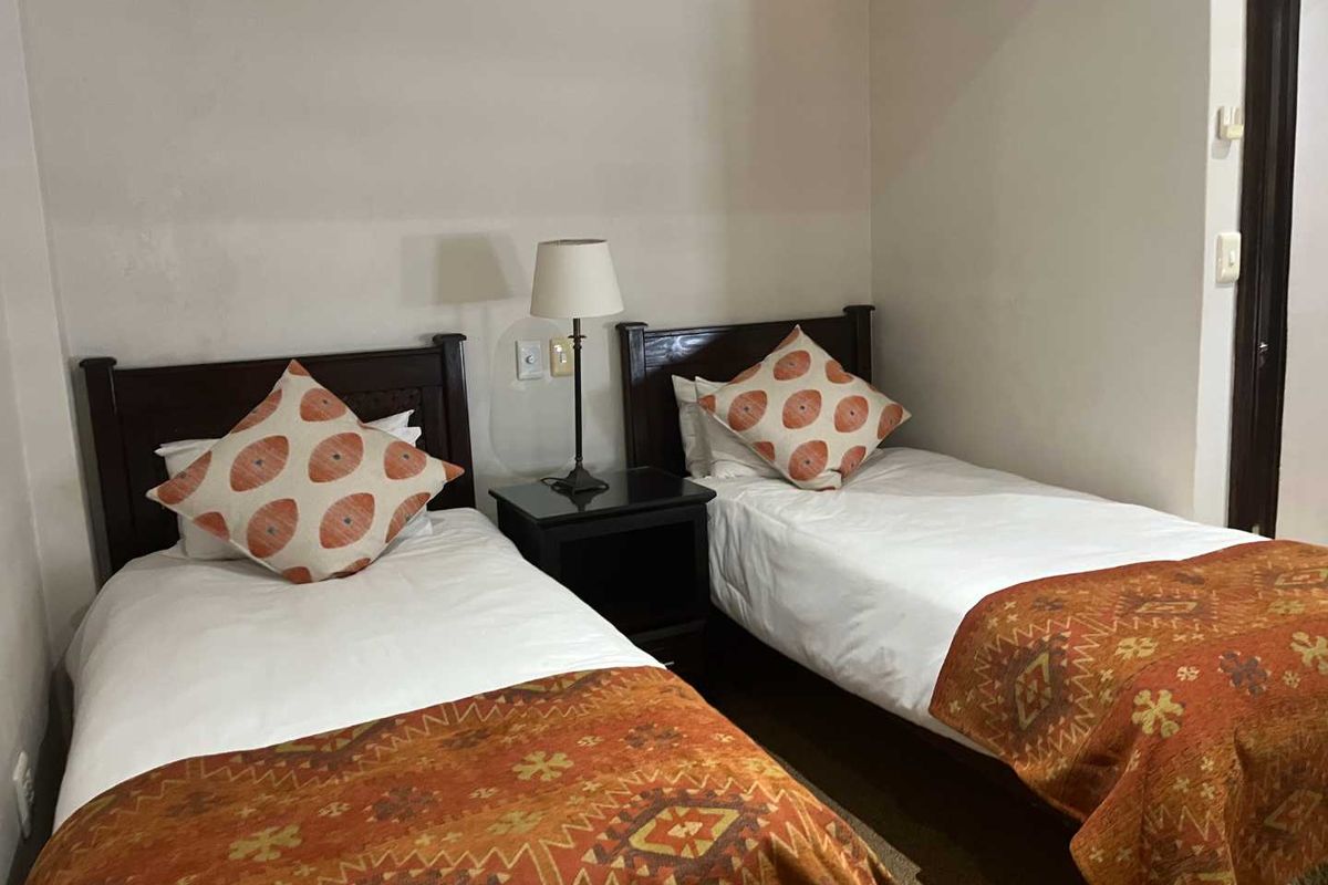 A bedroom featuring two single beds with a shared bedside table and lamp, finished with neutral walls and carpeted flooring.