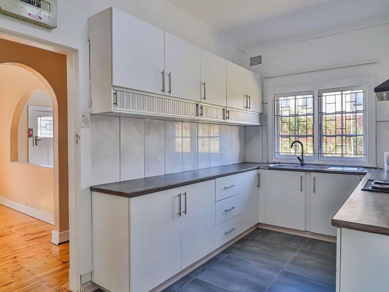 A clean, functional kitchen featuring white cabinetry, dark laminate countertops, a built-in hob with extractor, and tiled flooring, leading into an arched hallway.