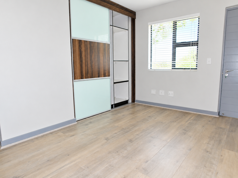 A bright, modern bedroom featuring light laminate flooring, a contemporary built-in wardrobe with sliding doors, and neutral wall finishes.