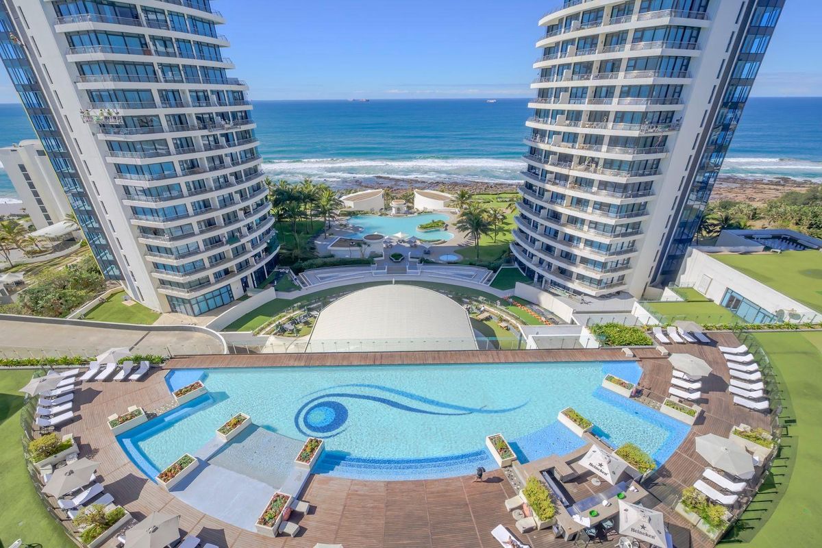 Aerial view of a luxury apartment complex featuring a large infinity swimming pool, timber decking, landscaped grounds, and direct ocean views.