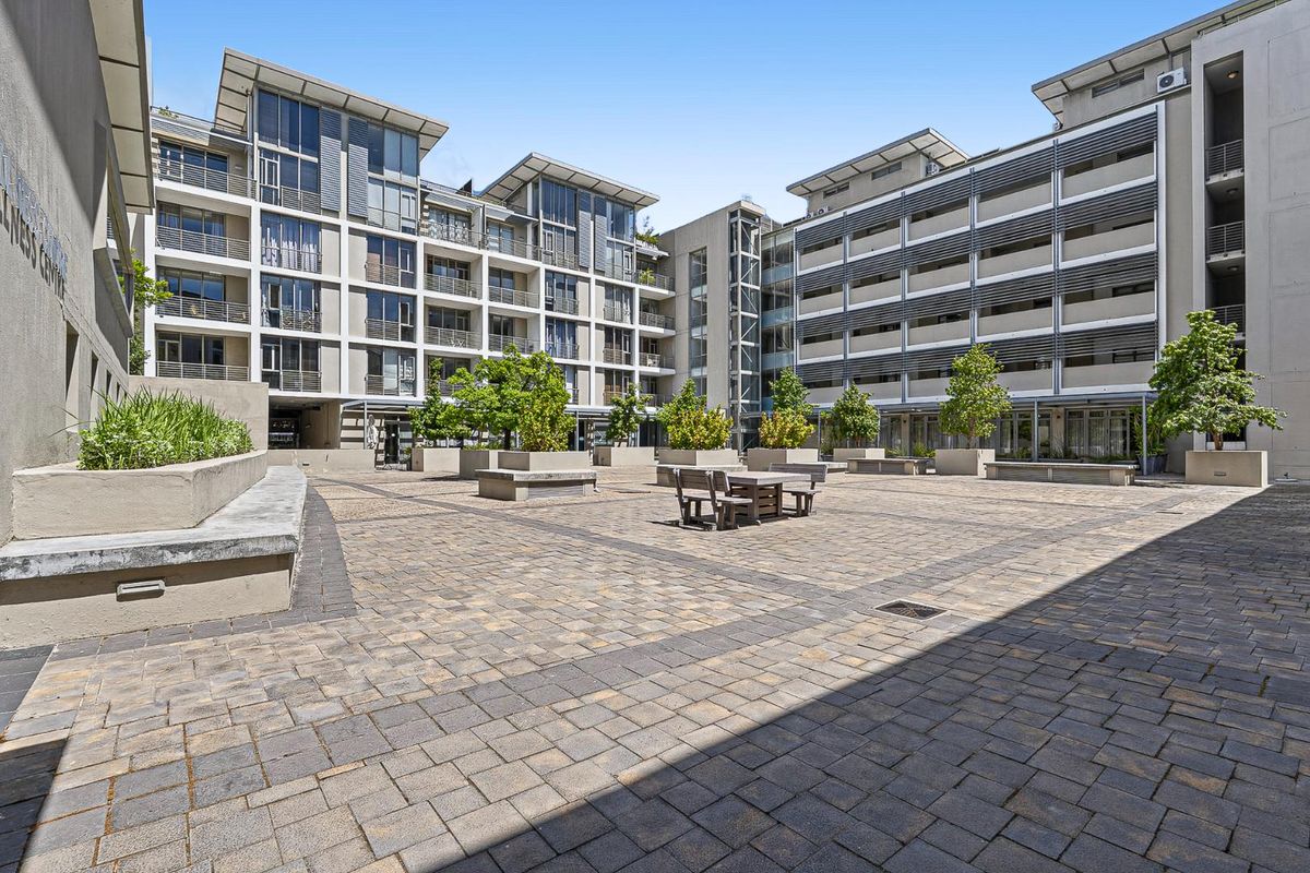 A spacious paved courtyard area within a modern apartment complex, featuring integrated concrete planters and multi-story building facades with balconies.
