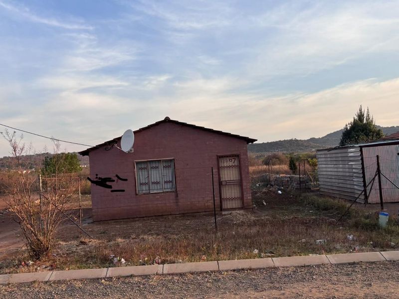 A detached single-story residential dwelling featuring a simple facade, security gate, and burglar bars, situated on an open plot with basic perimeter fencing.