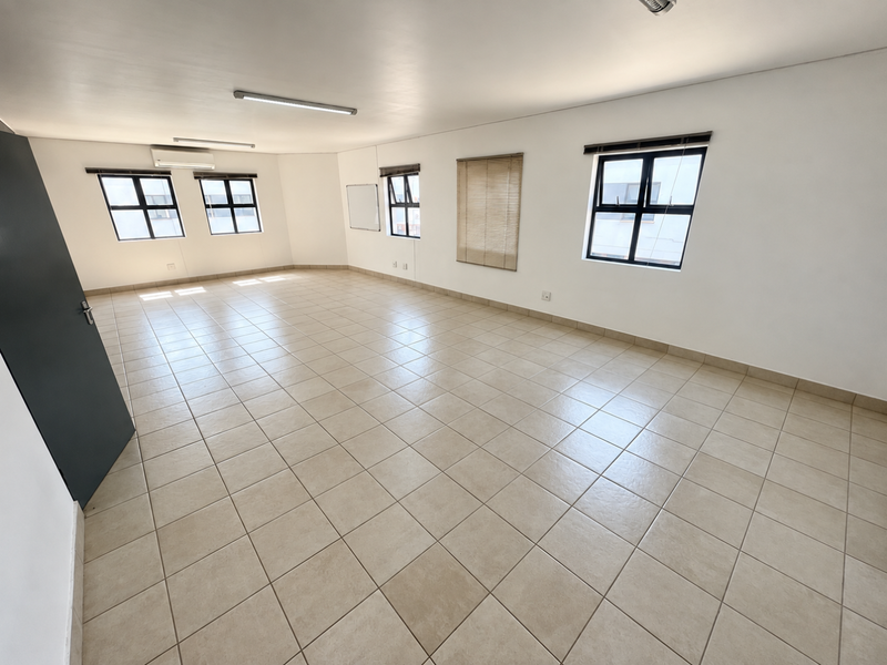 A spacious, empty office area featuring neutral ceramic floor tiles, multiple windows with blinds, and a wall-mounted air conditioning unit.