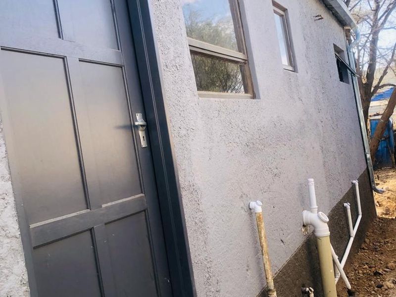 Side view of the property showing a textured plaster facade, dark grey door, aluminium windows, and exposed external plumbing infrastructure.