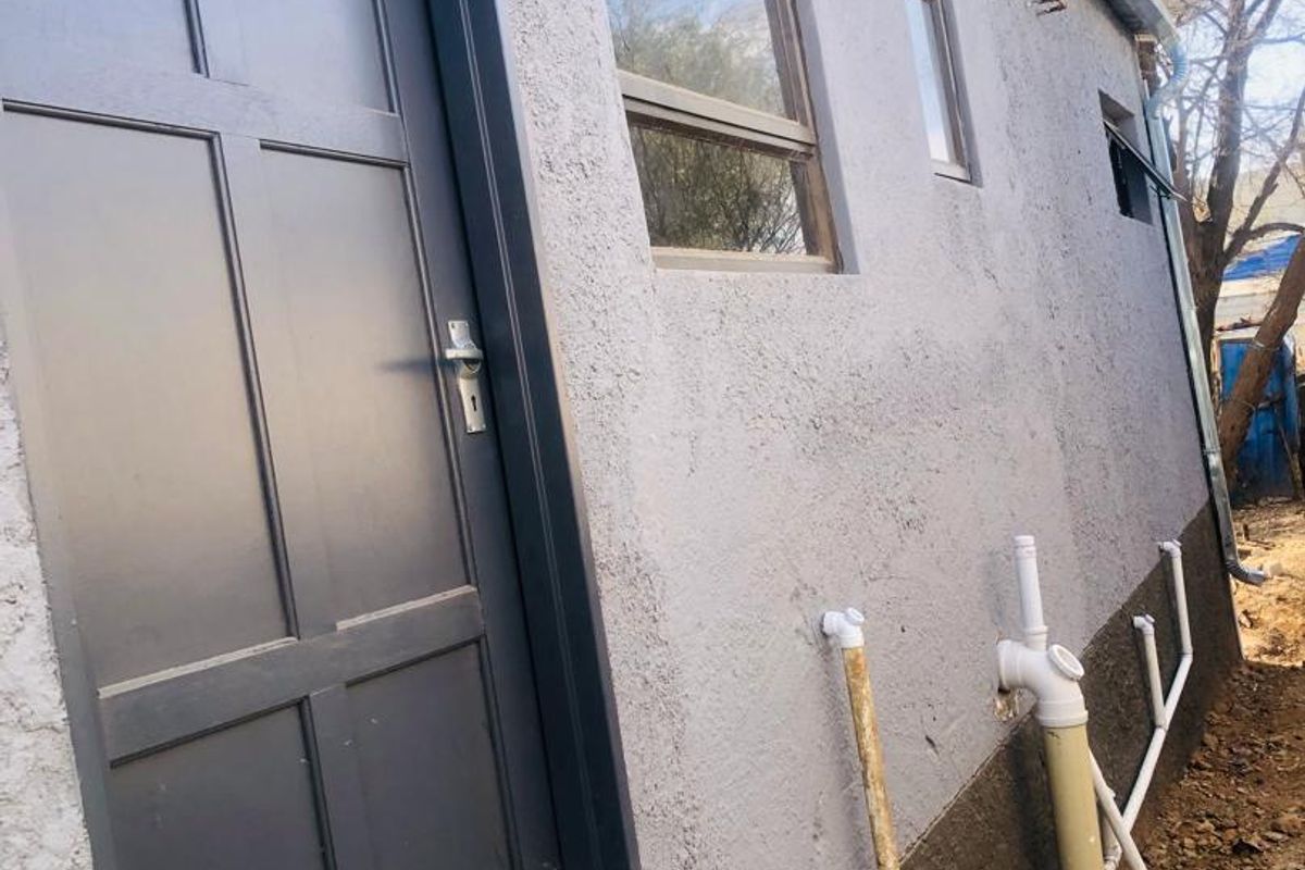 Side view of the property showing a textured plaster facade, dark grey door, aluminium windows, and exposed external plumbing infrastructure.