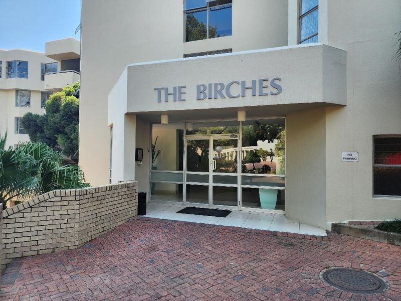 The front entrance of The Birches apartment complex, featuring a brick-paved driveway, a covered tiled porch, and clear glass entry doors.