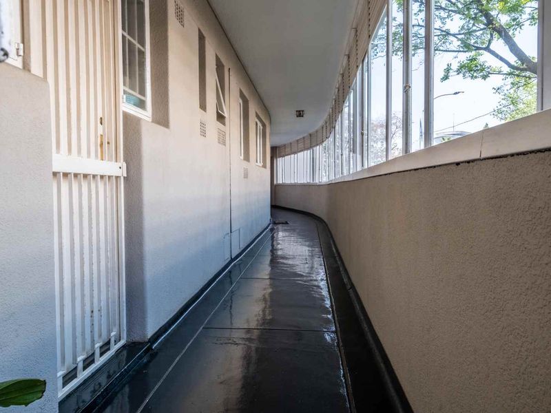 A long, curved apartment hallway featuring painted concrete floors, security gates, and large windows providing natural light and exterior views.