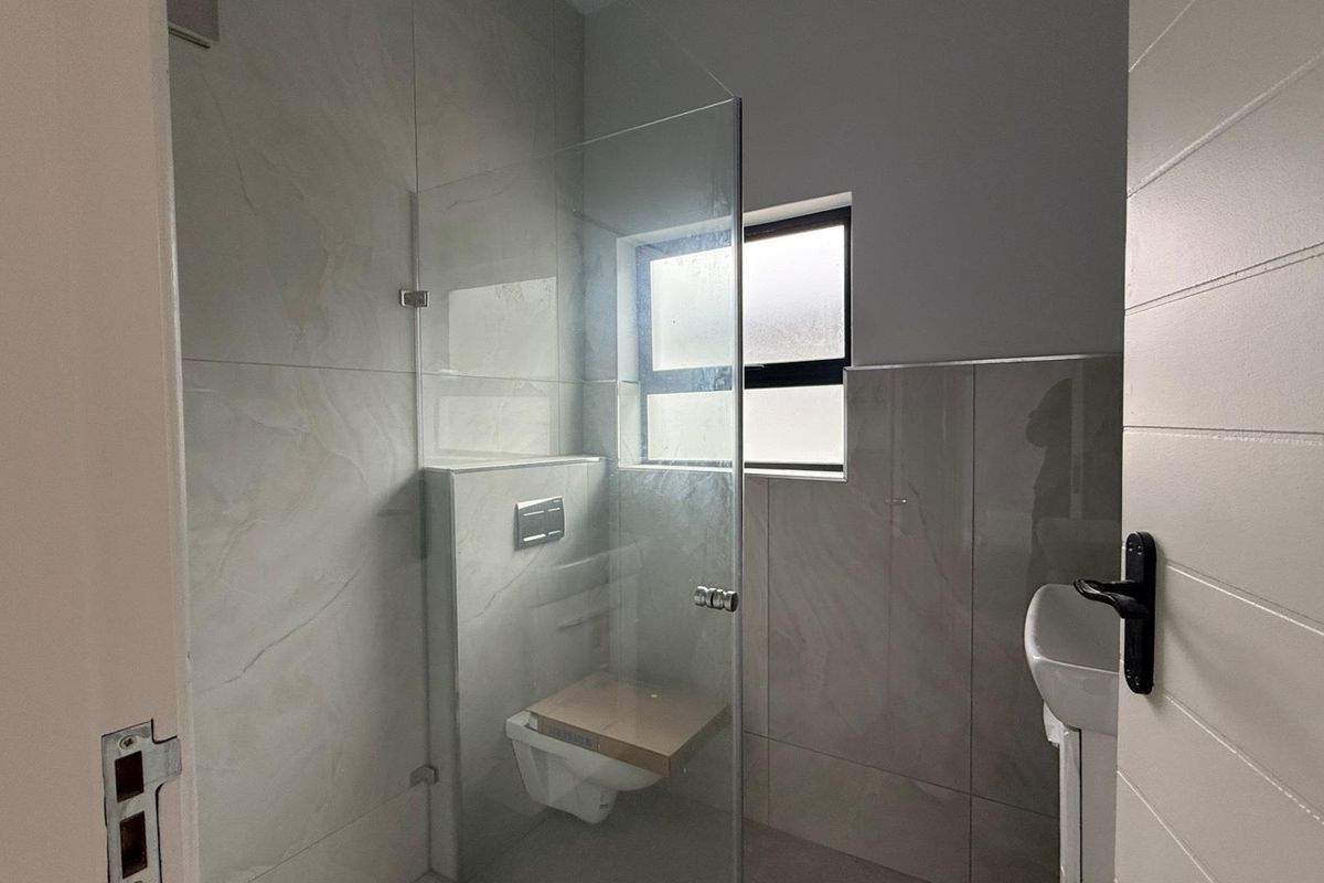 A modern, clean bathroom featuring a wall-mounted toilet, glass shower screen, and pedestal basin with neutral wall tiling.