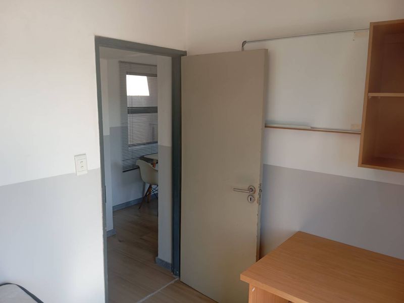 A simple bedroom featuring laminate flooring, built-in shelving, and a doorway leading to an adjacent room with window blinds.