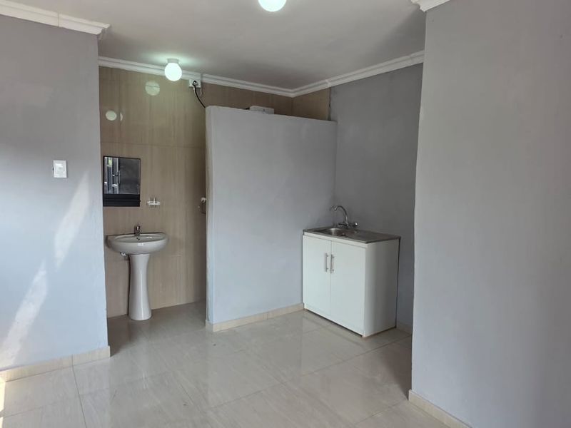 An open-plan studio space featuring tiled flooring, a pedestal basin, and a basic kitchen sink cabinet unit.