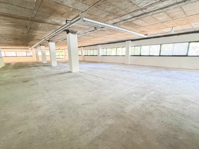 A large, open-plan commercial space featuring exposed concrete ceilings, industrial ducting, structural columns, and expansive windows providing ample natural light.