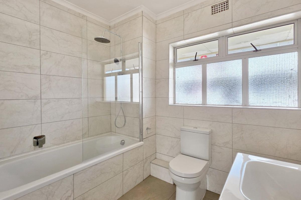 A clean, modern bathroom featuring a bathtub with a glass shower screen, a toilet, and a washbasin, finished with neutral marble-look wall tiles.