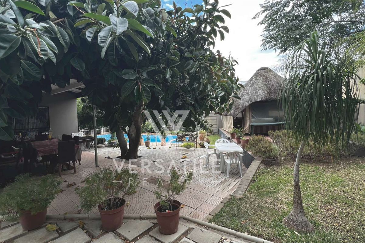 A spacious outdoor entertainment area featuring a paved patio, swimming pool, and a traditional thatched lapa structure set within a landscaped garden.