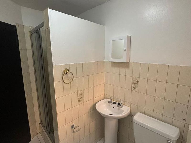 A functional bathroom featuring a pedestal basin, toilet, and a shower enclosure with tiled finishes, requiring cosmetic updates to modernize the space.