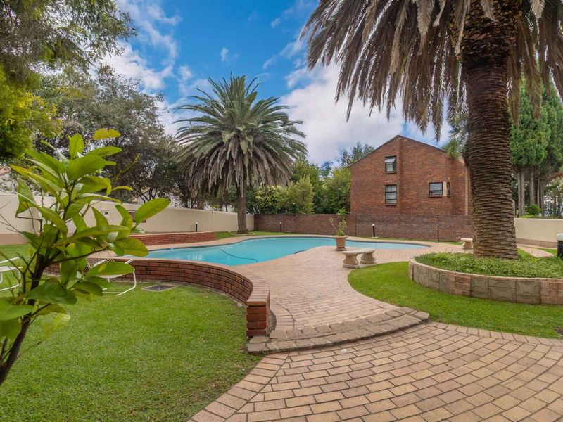 A well-maintained communal pool area featuring brick paving, landscaped garden surrounds, and established palm trees within a secure complex.