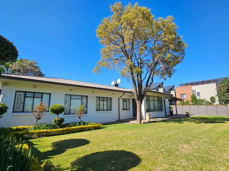 A well-maintained rear exterior view of a single-story brick house featuring a spacious lawn, mature tree, and established garden beds.