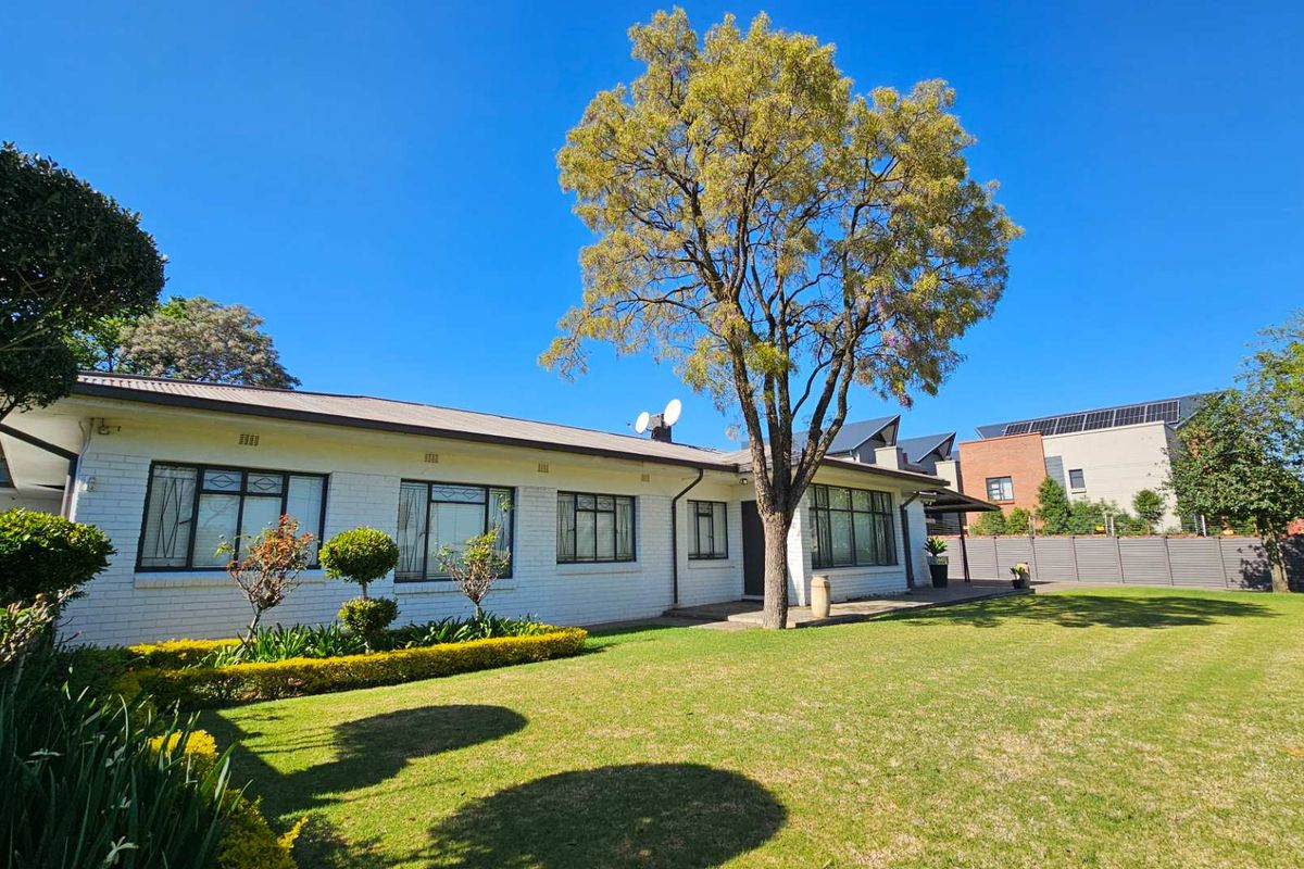 A well-maintained rear exterior view of a single-story brick house featuring a spacious lawn, mature tree, and established garden beds.