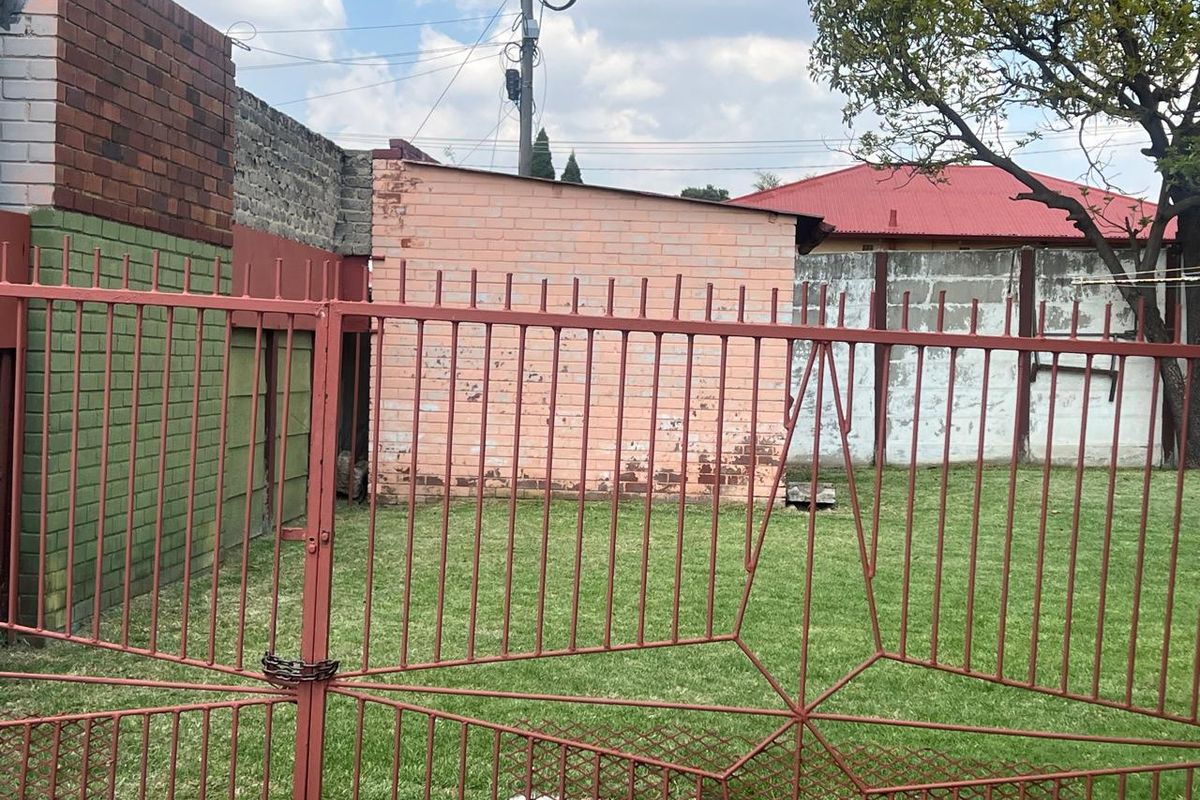 A rear garden area featuring a lawn, enclosed by a metal security gate and brick boundary walls with visible weathering.