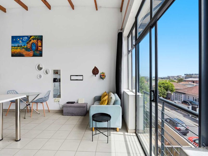 Bright open-plan living area featuring exposed timber ceiling beams, tiled flooring, and expansive sliding glass doors opening to a balcony with neighborhood views.