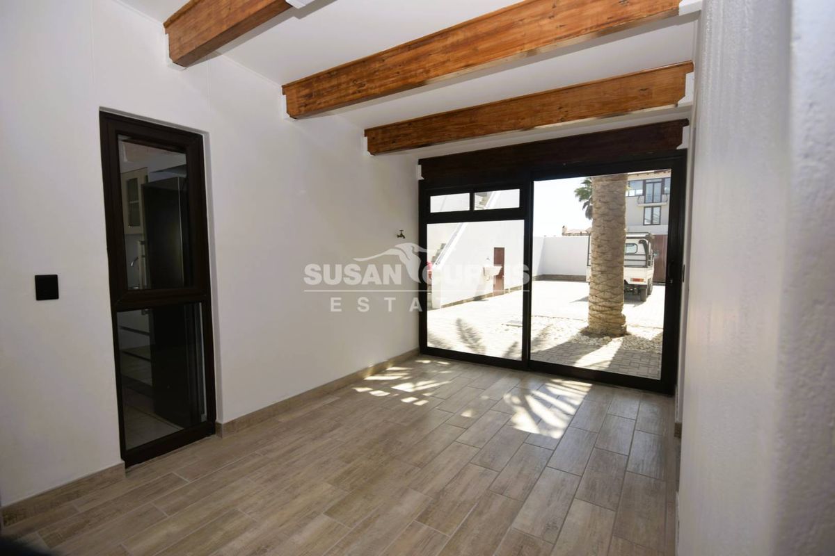 A bright, empty living area featuring exposed timber ceiling beams, wood-look tiled flooring, and large sliding glass doors opening to an exterior courtyard.