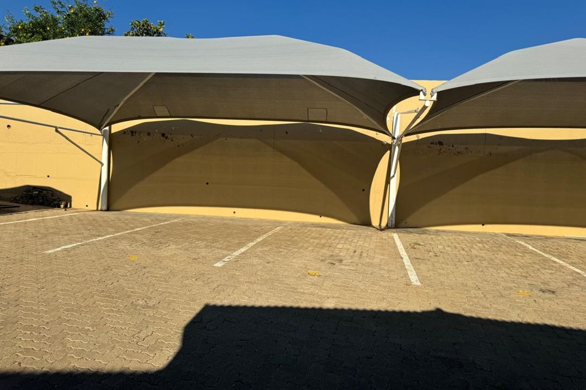 Outdoor parking area featuring paved ground and overhead shade sail structures against a retaining wall.