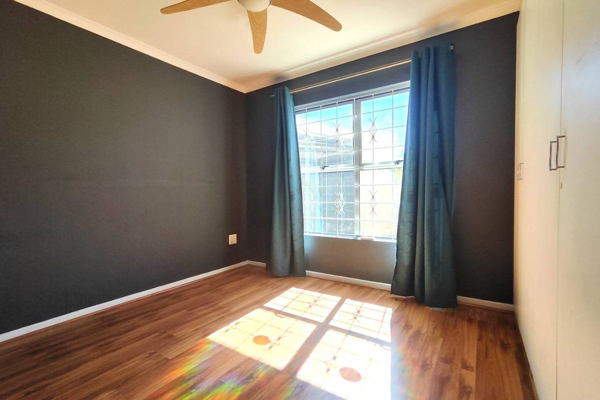 A bright, empty bedroom featuring dark accent walls, laminate flooring, built-in cupboards, and a ceiling fan, with security bars installed on the window.