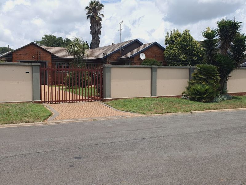 Front view of a brick house featuring a secure perimeter wall, metal driveway gate, paved entrance, and established garden.