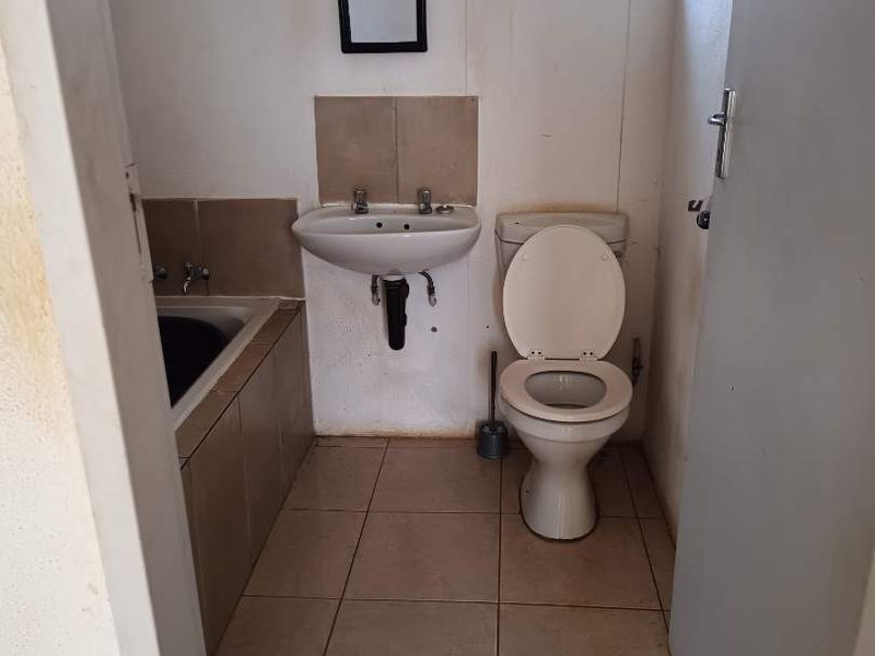 A compact bathroom featuring a built-in bathtub, wall-mounted basin, and toilet, finished with ceramic floor tiles and a window with security bars.