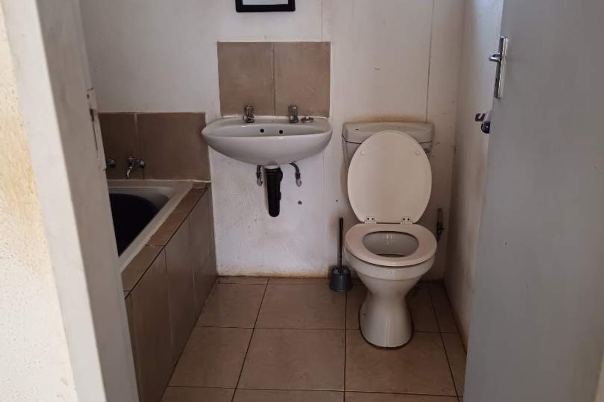 A compact bathroom featuring a built-in bathtub, wall-mounted basin, and toilet, finished with ceramic floor tiles and a window with security bars.