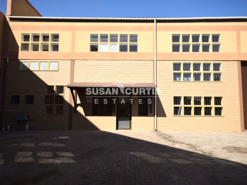 Front elevation of a multi-story industrial building featuring a paved entrance area, large multi-pane windows, and a covered entryway.