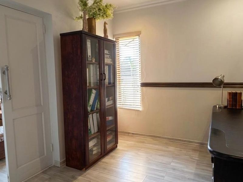 A functional home office featuring light-toned laminate flooring, a ceiling fan, and a window fitted with venetian blinds.