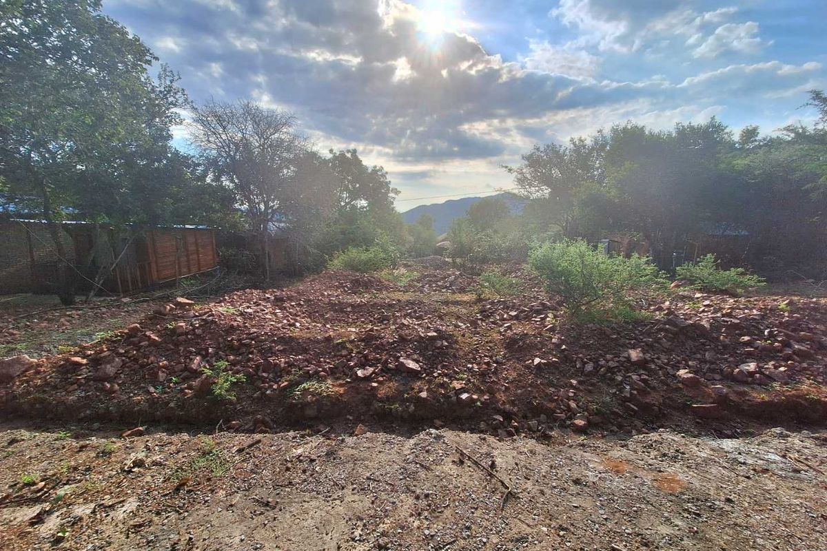 A view of a vacant residential plot featuring natural vegetation, mature trees, and uneven terrain with piles of excavated rock and soil.