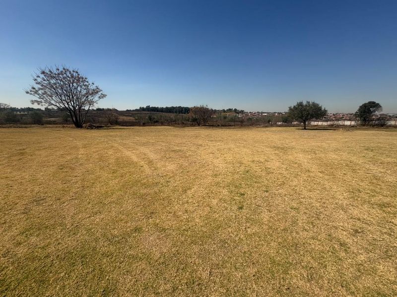 A wide, open residential plot featuring a flat grass lawn and mature trees, offering a clear view of the surrounding landscape.