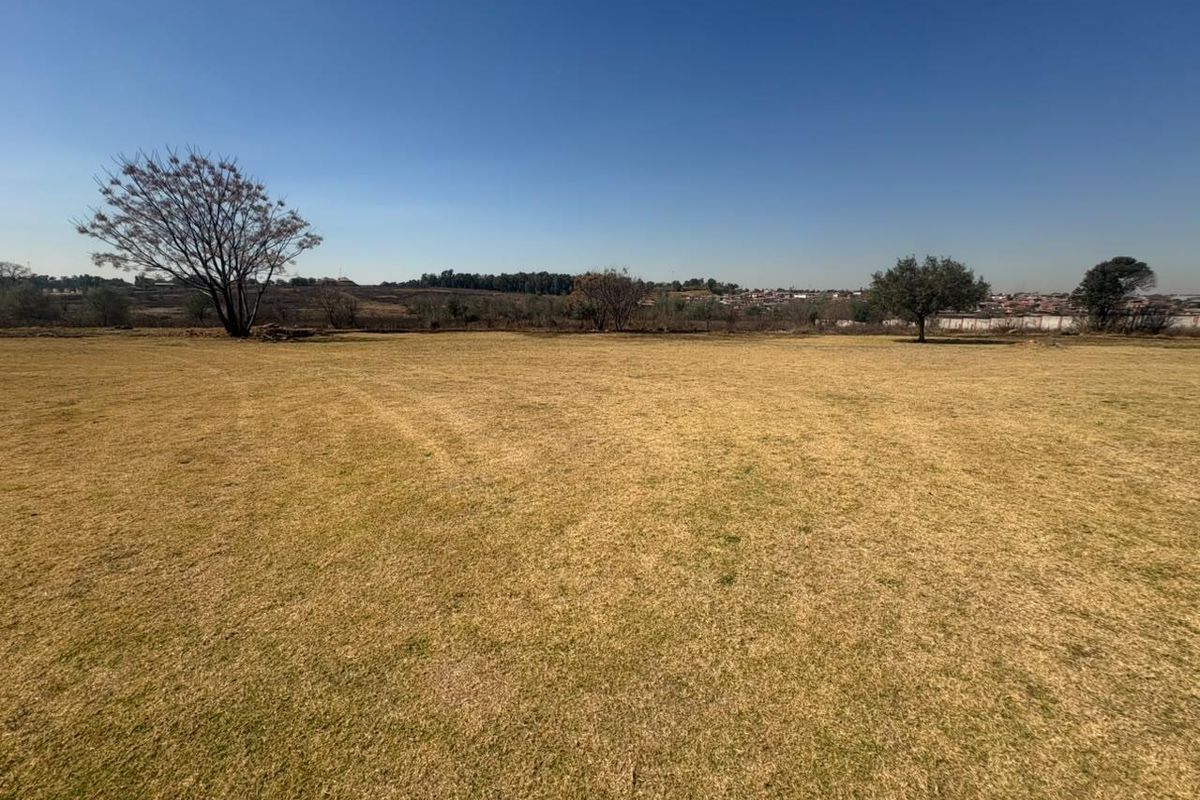A wide, open residential plot featuring a flat grass lawn and mature trees, offering a clear view of the surrounding landscape.