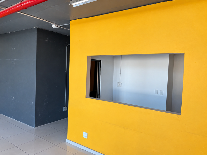 An open-plan commercial space featuring a bold yellow accent wall with a pass-through window, exposed industrial ceiling pipes, and neutral tiled flooring.