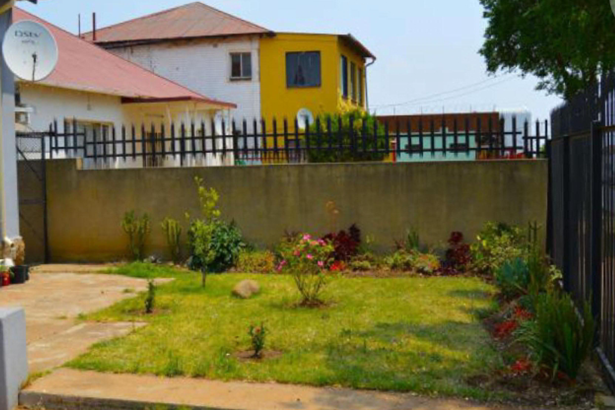 A modest rear garden area featuring a small lawn, landscaped garden beds, and concrete paving, enclosed by a perimeter wall and metal fencing.