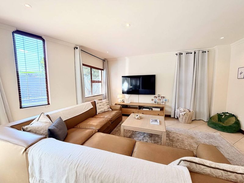 A bright and spacious living room featuring tiled flooring, recessed downlighting, and large windows with fitted blinds and curtains.