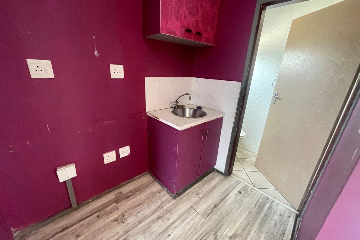 A compact kitchenette area featuring a small sink unit, overhead cabinetry, and direct access to an adjacent bathroom with tiled flooring.