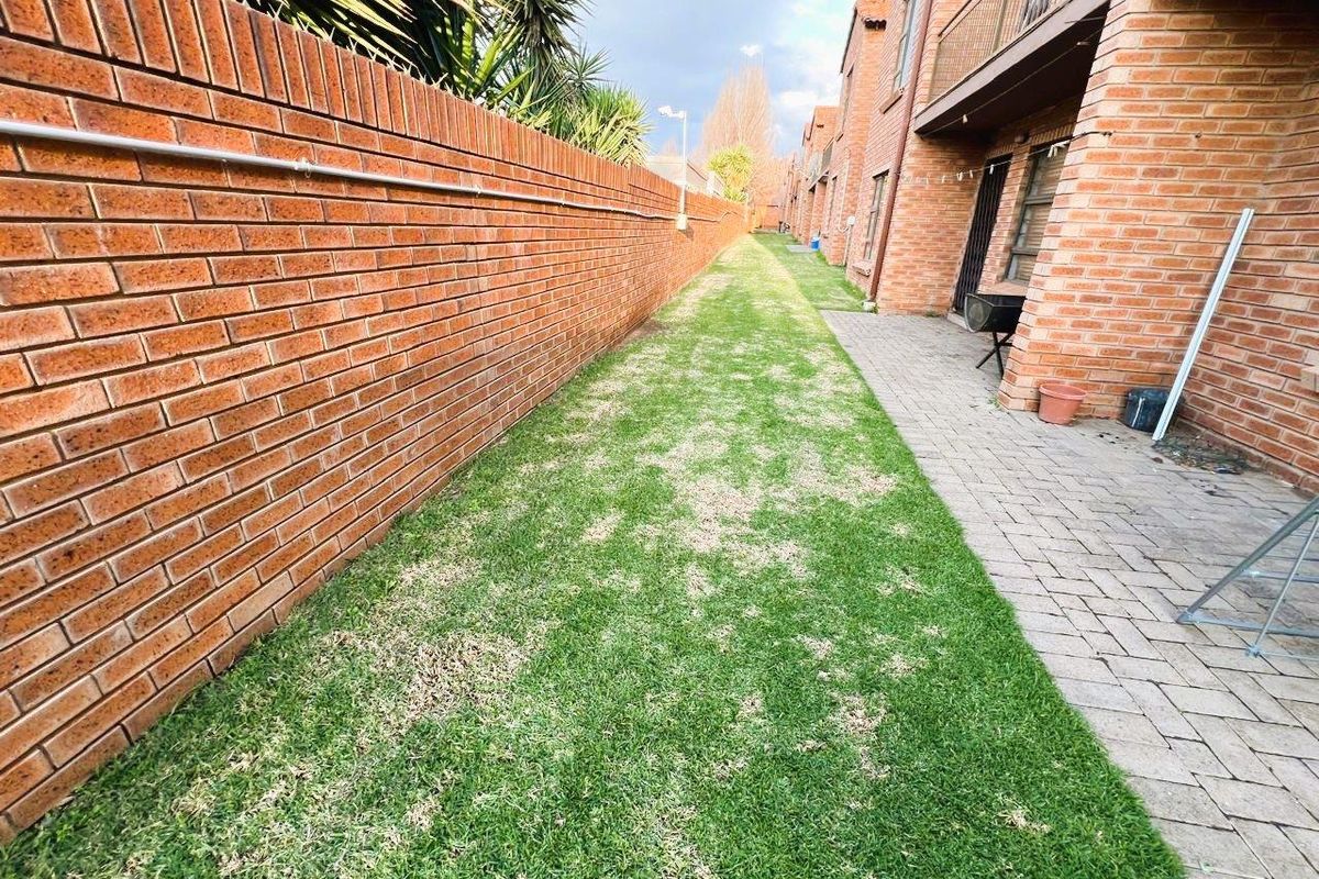 A rear exterior view of an apartment complex featuring a long brick boundary wall, a paved walkway, and a grass lawn area.