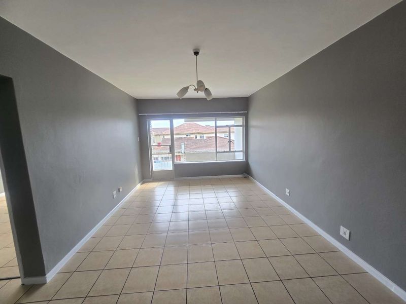 A spacious, empty living room featuring neutral grey walls, tiled flooring, and a large window providing natural light and access to an outdoor area.